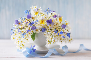 Still life with a bouquet of spring white flowers of bird cherry, blue pansies, scilla and brunnera in a cup on a table