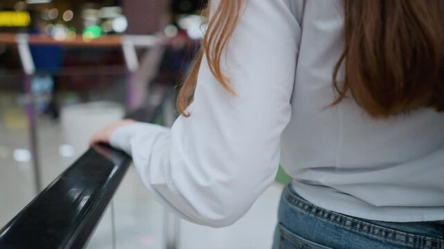 Rear view of female shopper stepping off moving escalator in bustling mall, holding shopping bags, she wears casual attire with phone tucked in back pocket, navigating modern retail environment