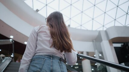 Back view of young lady on moving escalator in modern mall, phone in back pocket, hand on rail, with other people ascending in front, showcasing everyday shopping experience and urban lifestyle