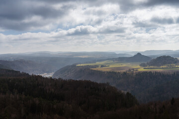 Obraz premium Panoramic view of forested canyon and towering sandstone cliffs under overcast skies in Saxon Switzerland, showcasing the dramatic Schrammsteine rock formations.