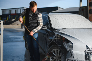 Naklejka premium Car wash equipment. A man is washing a car at self service car wash. High pressure vehicle washer machine sprays foam