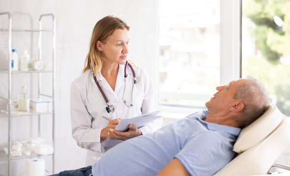 Positive female doctor conducting questionnaire of elderly male patient lying on couch in medical office, asking series of questions and noting down responses in document folder - Powered by Adobe
