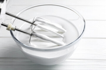 Mixing whipped cream with hand mixer in bowl on white wooden table, closeup