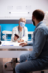 Obraz premium Elderly female doctor sits with bearded man and advises him on medication use for treatment. Senior physician giving bottle of prescribed pills to male patient, both wearing face masks in clinic room.
