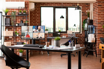 Empty office desk with computer screen displaying group virtual meeting in progress. Videoconference call of caucasian people on desktop monitor in spacious startup workspace with modern furniture.