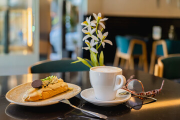 Cappuccino with creamy foam and elegant eclair on black cafe table with soft daylight. Concept of cozy lifestyle, delicious dessert break and calm morning in modern urban coffee shop