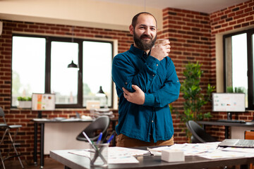 Fototapeta premium Young businessman holding coffee cup and smiling towards camera prepared to work startup office. Portrait of male analyst standing in brick wall workspace surrounded by company research documents.