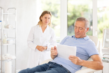 Before medical manipulation, mature man patient sits in chair and reads documents with woman doctor...