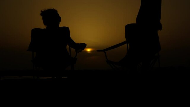 Parting at sunset. Silhouette of a man walking away while a woman sits alone, gazing at a stunning sunset from beach chairs. The scene captures a moment of emotional distance and heartbreak