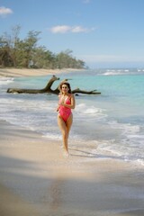 Fototapeta premium A confident young woman walks gracefully on a picturesque Hawaii beach in a stylish bikini, surrounded by greenery and the ocean sounds, enjoying the warm sunshine and embodying summers essence