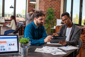 Caucasian man and black coworker analyze financial reports using tablet and documents, discussing strategy ideas. Business partners use digital device in planning and collaboration for company growth.