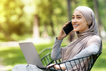 Young woman in hijab talking on phone and using laptop while sitting on bench at park, working outdoors, free space