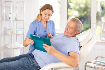 Obraz premium Female doctor obtaining consent of patient for medical procedures or therapies. Focused senior man reading and signing medical agreement while lying on couch