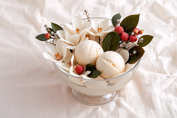 Creamy ice cream in a glass bowl is beautifully decorated with white blossoms, green leaves, and red berries, all resting on a soft white cloth.