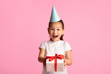 Holiday Present. Excited asian girl wearing party hat and holding gift box, isolated on pastel pink studio wall, copyspace