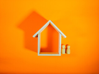 A couple of peg doll stand near a wooden house on a orange background. A young family is standing near their house. The concept of buying a home. The property. Family happiness. Unit of society.