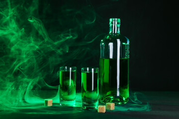Absinthe in shot glasses, bottle and brown sugar on table against black background with smoke