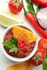 Spicy salsa sauce in bowl, nachos and ingredients on white table, flat lay