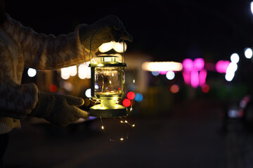 Woman holding Christmas lantern with fairy lights in darkness, closeup. Bokeh effect