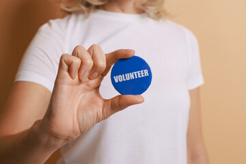 Woman holding button badge with word Volunteer on beige background, closeup