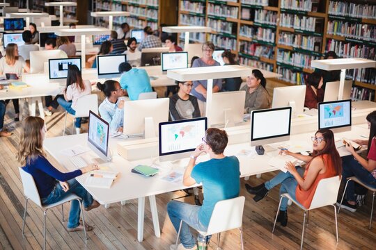 Students in a library using computers. Diverse group studying, collaborating, and learning. Busy library with technology and books. Academic environment. International students working on computers.