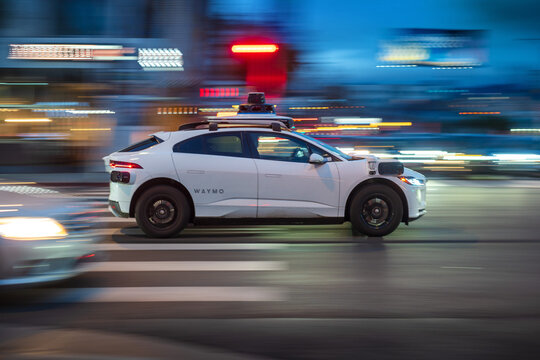 LOS ANGELES, CA - March 14, 2025: Google's Alphabet Waymo driverless self driving taxi cab rushing through city traffic, crossing illuminated street intersection at dusk in West Hollywood, California.
