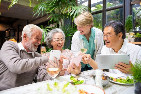 Group of elderly friends enjoying lunch, laughing, and using smartphones and tablets. Diverse seniors sharing joyful moments and technology at a restaurant. Happy diverse seniors on social media. - Powered by Adobe