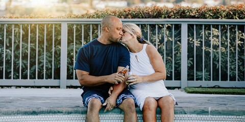 Couple sitting by a pool, enjoying a moment together. They are sharing a kiss, holding drinks, and relaxing by the poolside, enjoying the sunny day. Diverse couple kissing while sitting by pool.