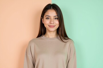 Obraz premium A Smiling Young Woman with Long Hair Wearing a Casual Beige Sweater, Posed Against a Colorful Pastel Background in Soft Peach and Mint Green Tones