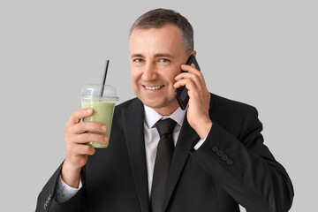 Mature businessman with glass of vegetable smoothie talking by mobile phone on light background
