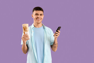Handsome young man with tasty doner kebab and mobile phone on lilac background