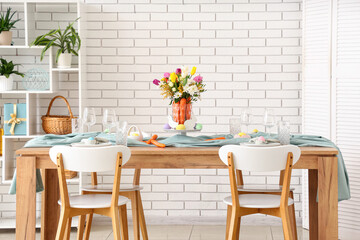 Beautiful table setting for Easter decorated with bouquet of carrots and flowers near white brick wall