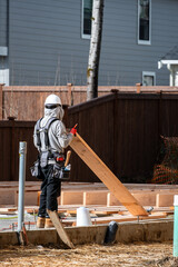 Obraz premium Construction worker, carpenter, in tool belt and hard hat on housing development project, installing subfloor wood on cement foundation, stacks of lumber, sunny winter day 