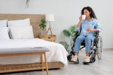 Young African-American woman in wheelchair with cup of coffee at home