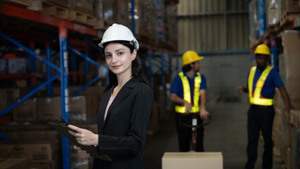 A woman wearing a hard hat and a suit stands in front of a warehouse with a clipboard in her hand