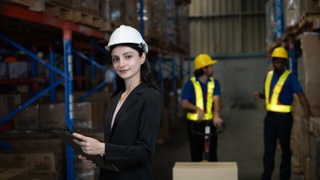 A woman wearing a hard hat and a suit stands in front of a warehouse with a clipboard in her hand - Powered by Adobe