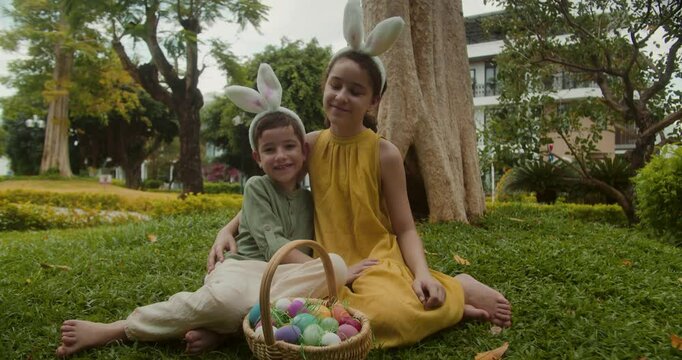 Children with bunny ear headbands on their heads are sitting in a meadow on Easter, hugging each other, looking at the camera, and smiling. Easter Egg Hunt. Kids collects Easter eggs