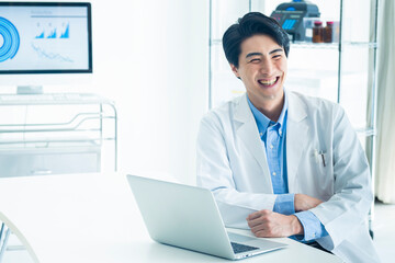 A cheerful male doctor in a white coat sits at a desk with a laptop in a bright hospital office. He is smiling, suggesting a positive work environment and friendly healthcare service.
