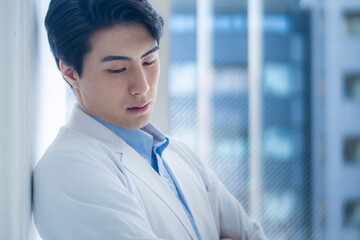 A young male doctor in a white lab coat stands in front of a hospital window, leaning against the wall with a pensive, downward gaze. He seems deep in thought or concerned.