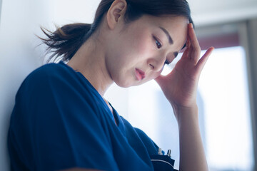 A close-up of a nurse in blue scrubs holding her forehead, eyes closed, showing signs of mental fatigue or stress in a clinical environment.