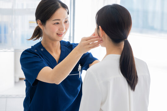 A doctor in blue scrubs gently examines a female patient's neck in a brightly lit medical clinic.