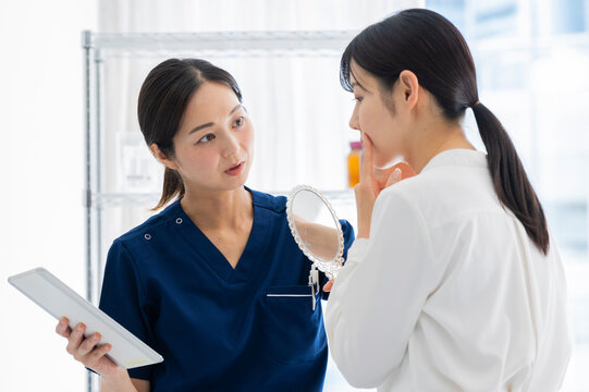 A female doctor in blue scrubs holds a mirror while consulting a female patient about her facial skin or features in a clinical setting.

