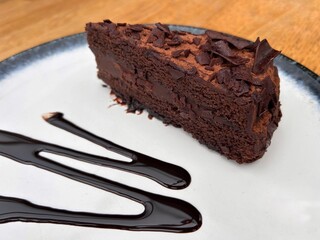 Close-up of a dark chocolate cake slice topped with flakes and cocoa powder, served on a white plate with a swirl of chocolate syrup
