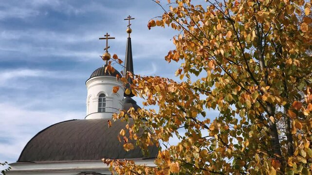 Yellow-orange autumn foliage swaying gently, partially veiling white-gray church with dark dome and golden crosses, standing against cloudy blue sky backdrop