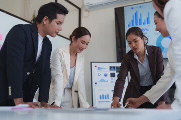 Asian businesspeople leaning over a table analyzing data on large paper sheets, with charts and graphs displayed on screens in the background, working together in a modern office