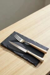 Fototapeta premium Elegant place setting featuring a fork and knife on a dark slate rectangular serving piece atop a light wood surface