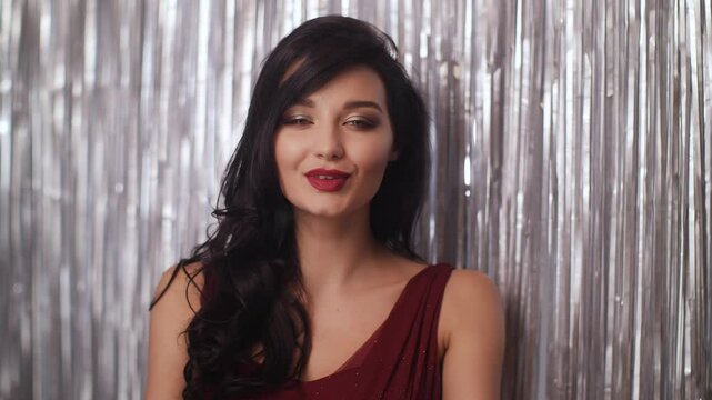 Beautiful brunette woman in a red dress blows a kiss in front of shimmering silver curtains. Conceptual promo footage ideal for romance, celebration, beauty or fashion ads.