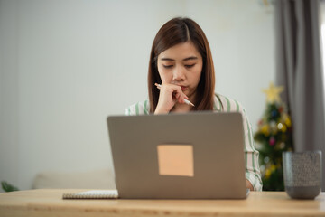 Asian Young woman thinking deeply while working on laptop at home during holiday season with festive decorations in the background
