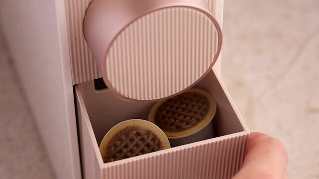 A woman's hand shows the compartment for used capsules from a capsule coffee machine in pale beige, champagne or rose gold. Recyclable plastic coffee capsules with a paper lid