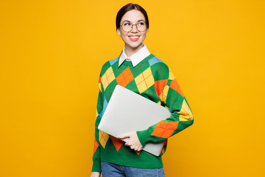 Side view young IT woman wear glasses green sweater casual clothes hold use closed laptop pc computer look aside on area isolated on plain orange yellow background studio portrait. Lifestyle concept.